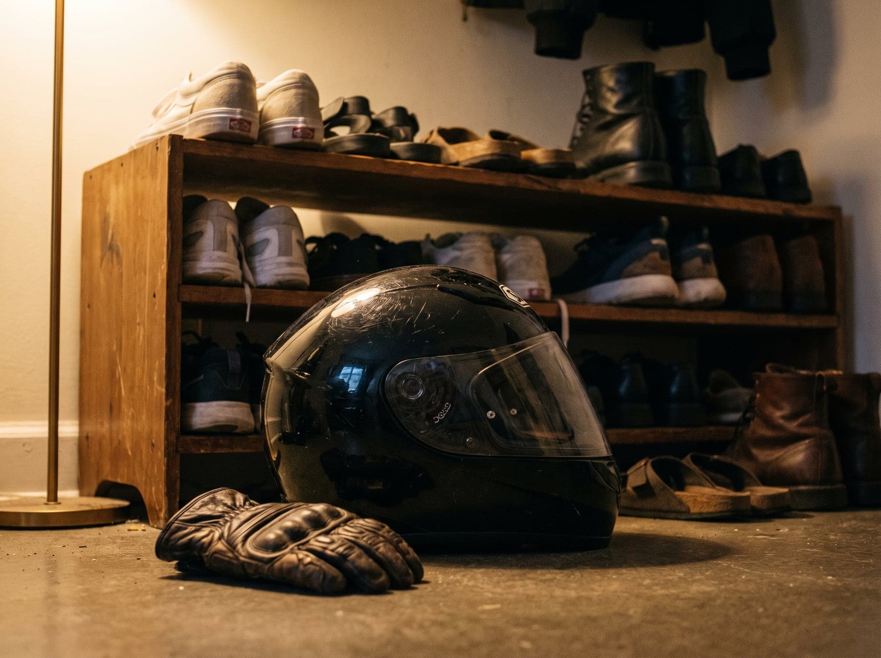 A scratched full-face helmet left on the floor by a shoe rack, a riding glove dropped beside it